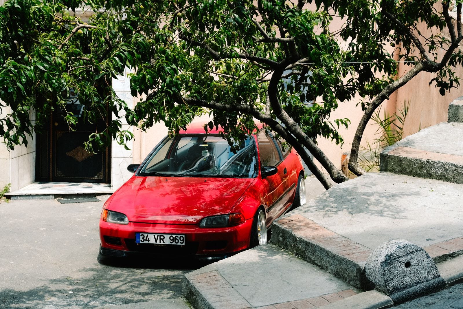 Rød hatchback parkeret i skyggen under et træ